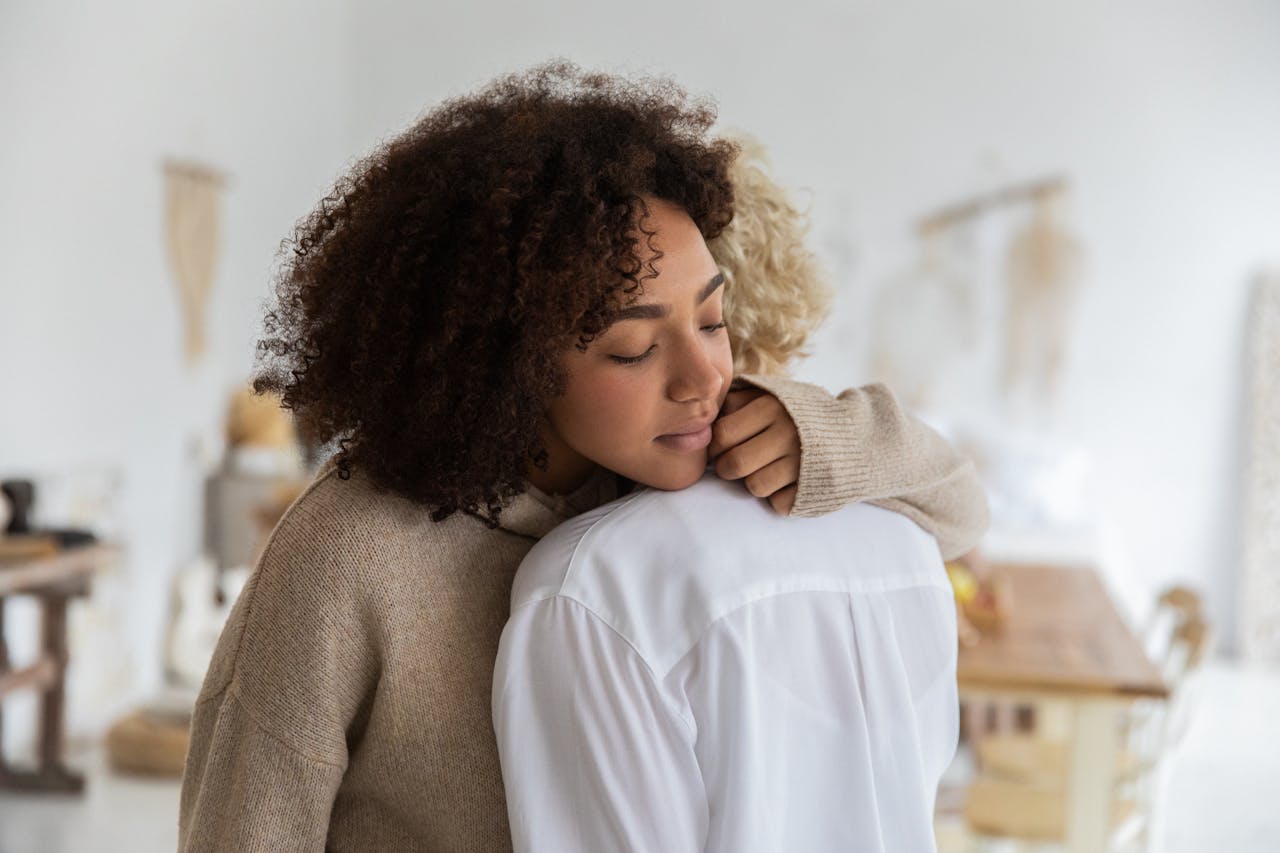African American woman embracing female anonymous friend while standing in cozy light room with wooden table and decorations on wall with blurred background