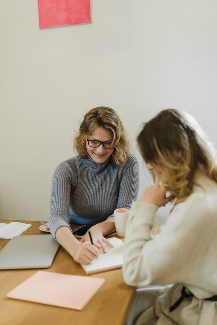 Two women in sweaters planning together at a wooden table with laptop and notebook.